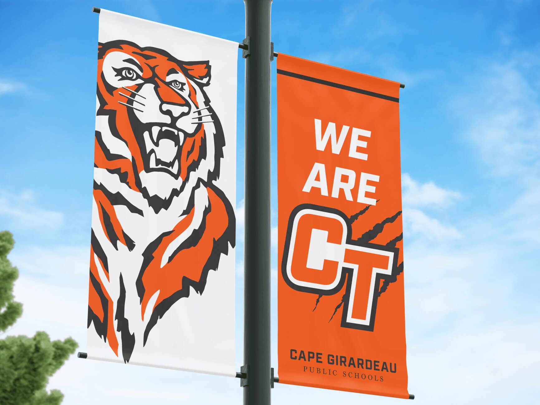 Two banners on a pole: white with orange tiger head, orange with 'WE ARE CT' and Cape Girardeau Public Schools, blue sky.