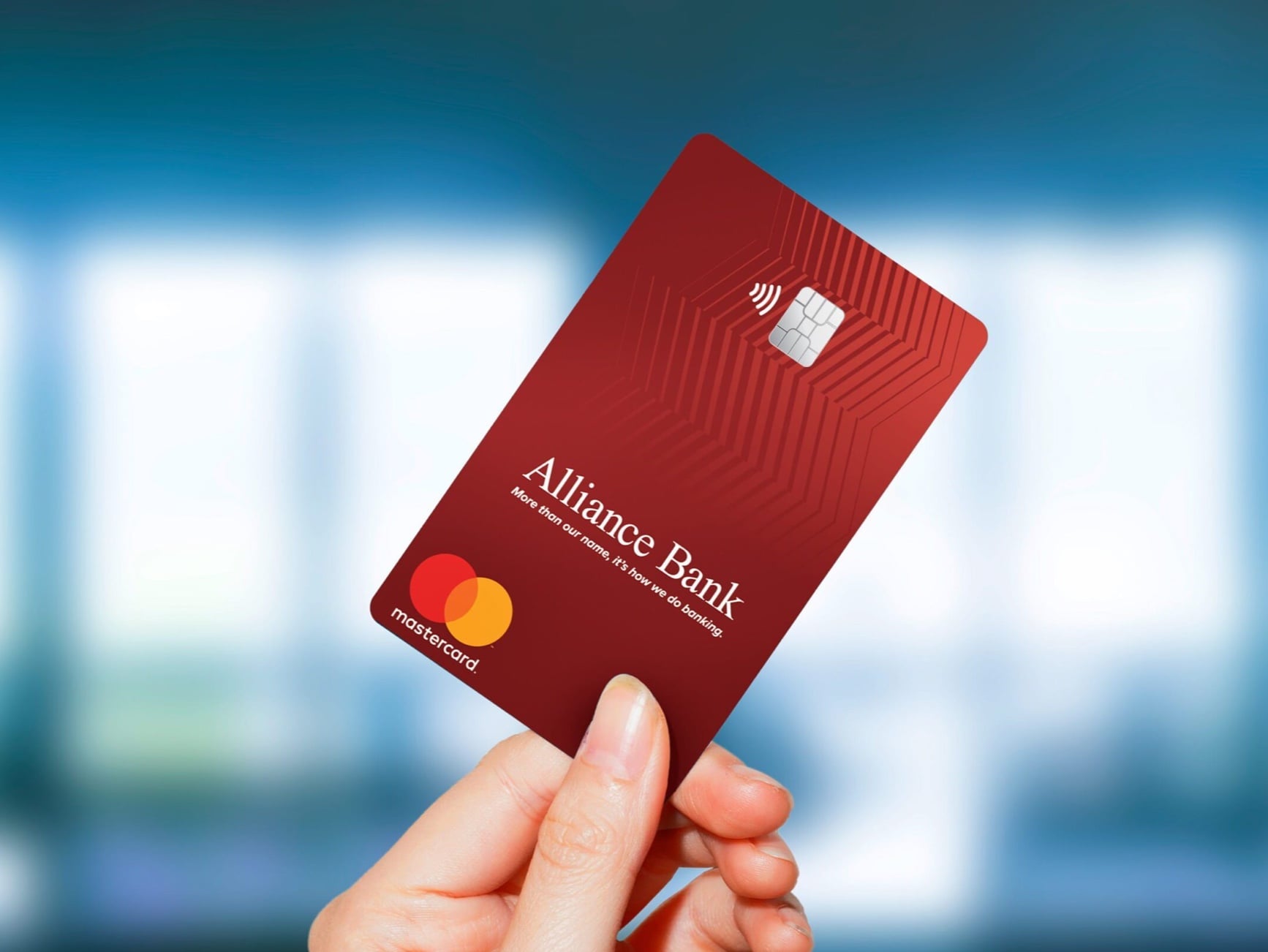 A hand holds a red Alliance Bank Mastercard credit card with chip and contactless symbol against a blurred blue background.
