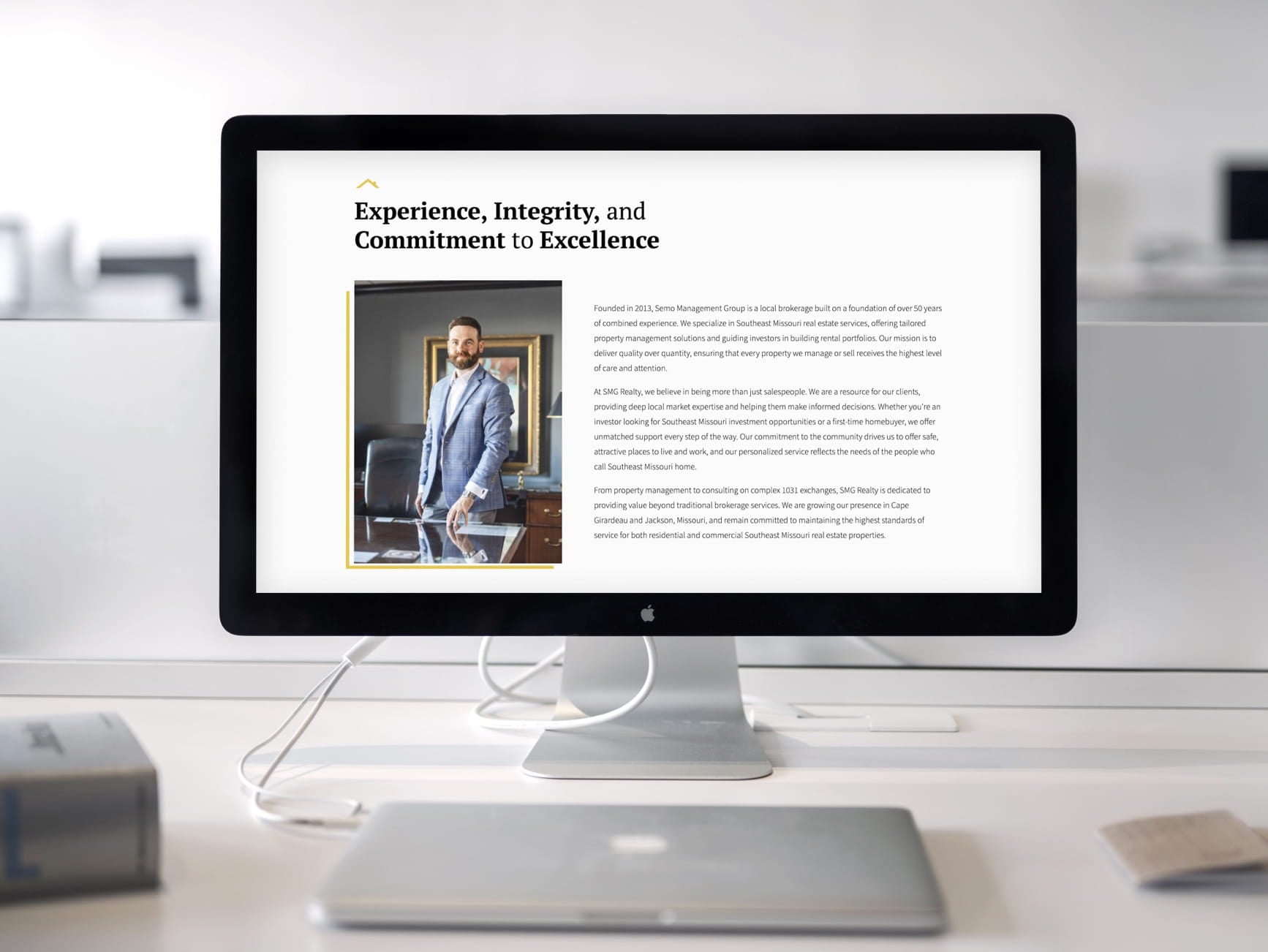 Apple iMac shows SMG Realty webpage with a man in a blue suit and text about experience, integrity, commitment.