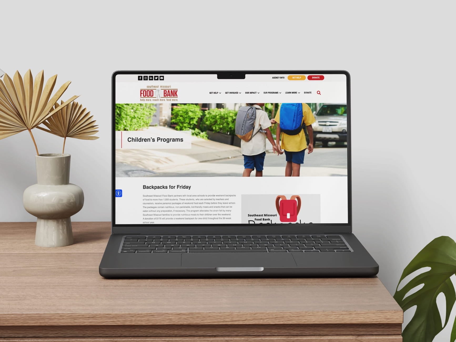 Laptop on desk shows Southeast Missouri Food Bank site with kids wearing backpacks, decorative plant vase with dried leaves.