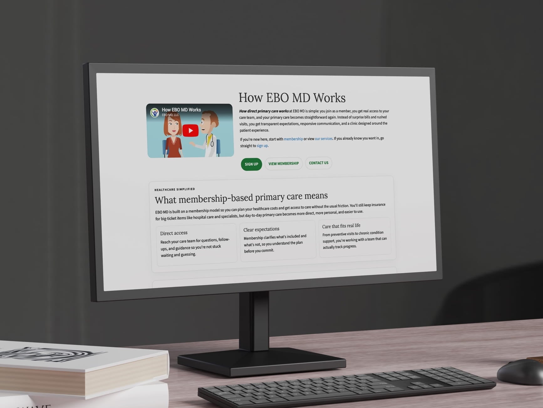 Monitor shows EBO MD site: 'How EBO MD Works' heading, video thumbnail, sign‑up buttons, membership info, desk with keyboar