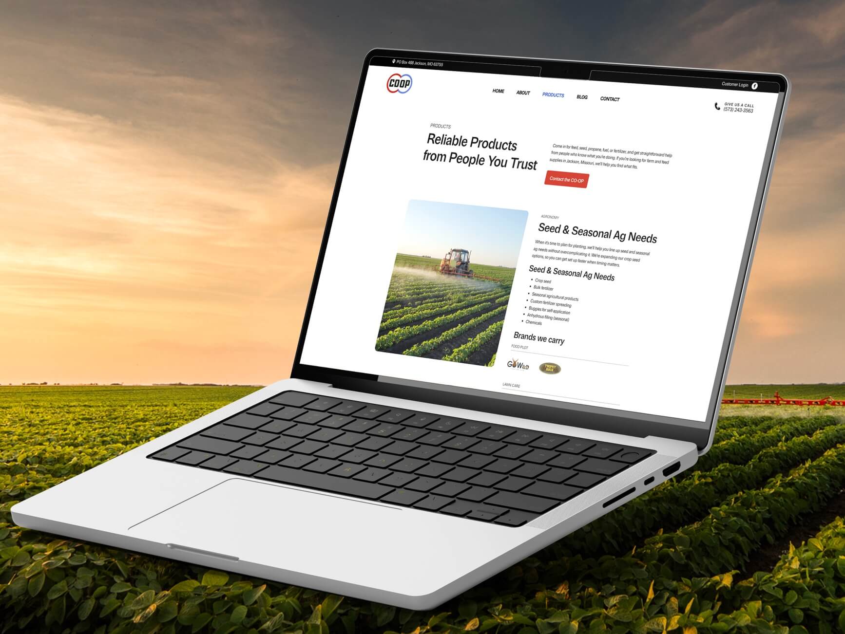 Laptop showing CO-OP ag website with seed and seasonal product list, set in a green field at sunset.