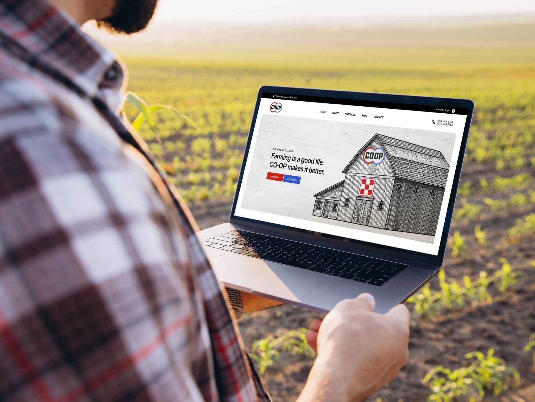 Man in plaid shirt holds laptop displaying CO-OP website with barn illustration over green field background.