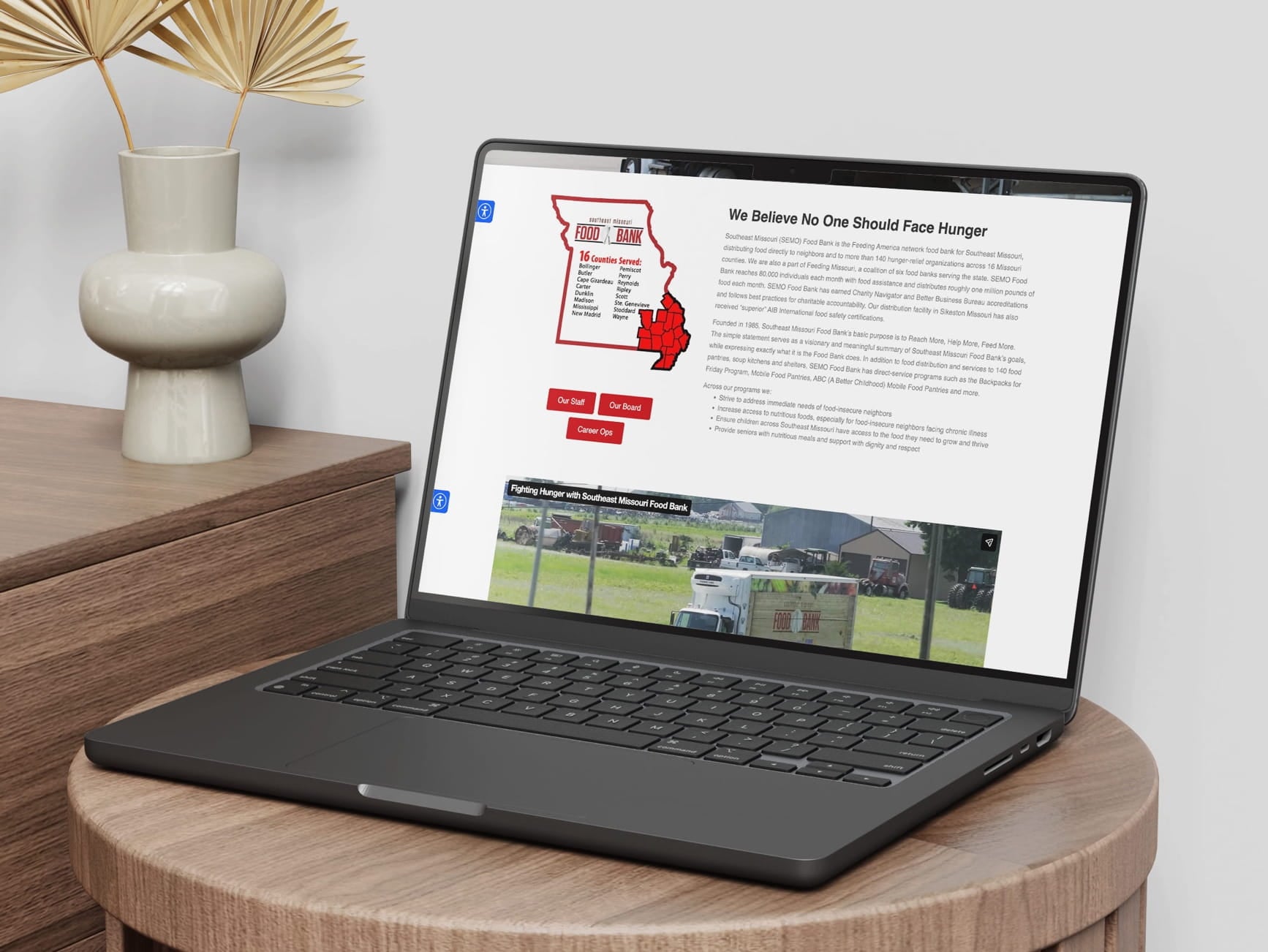 Laptop on a wooden table shows Southeast Missouri Food Bank website with map and text, next to a vase with dried palm leaves.
