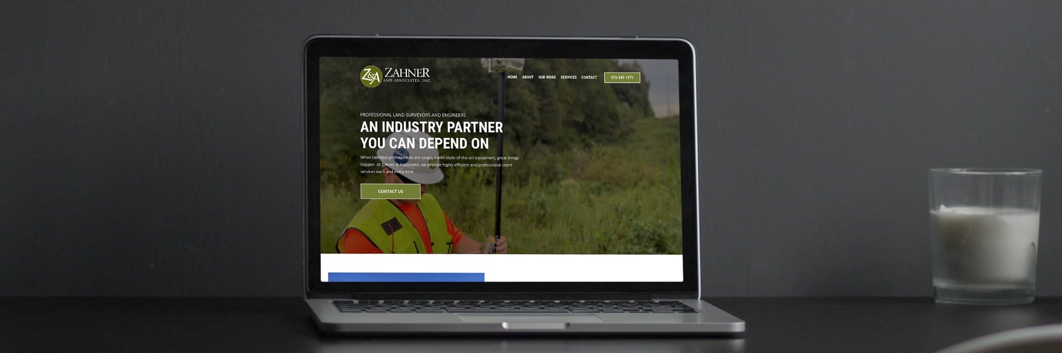 Laptop showing Zahner and Associates website with a surveyor in a safety vest, navigation menu, and contact button.