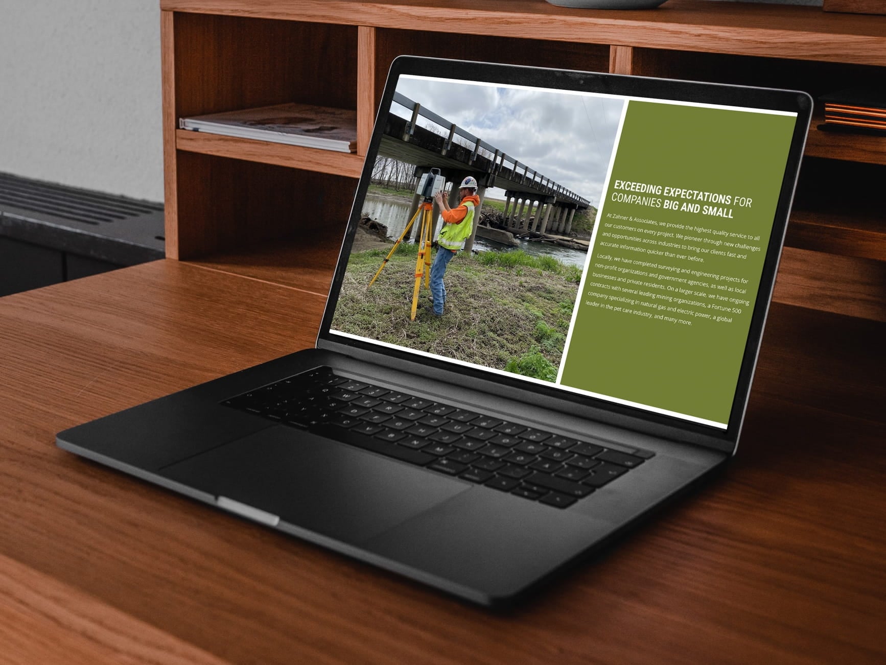 Laptop on wooden desk showing a webpage with a surveyor using a tripod instrument near a bridge