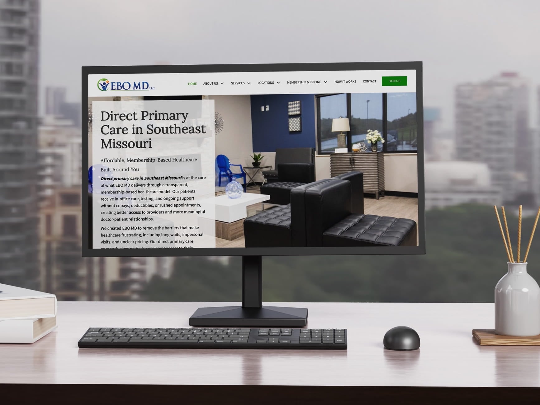 Monitor shows EBO MD direct primary care site, modern office photo, keyboard, mouse, and desk items.
