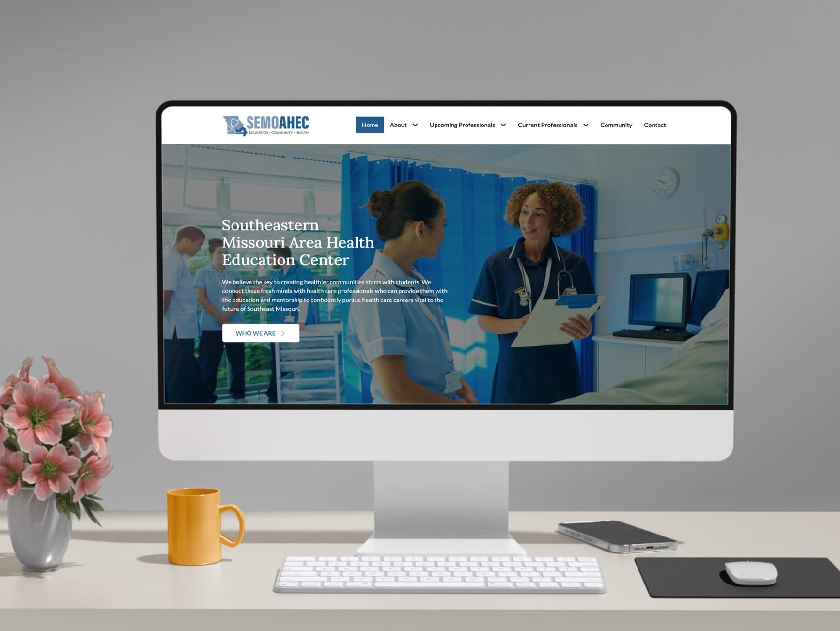Monitor displays health education site with nurses; desk holds yellow mug, pink flowers, keyboard, and phone.