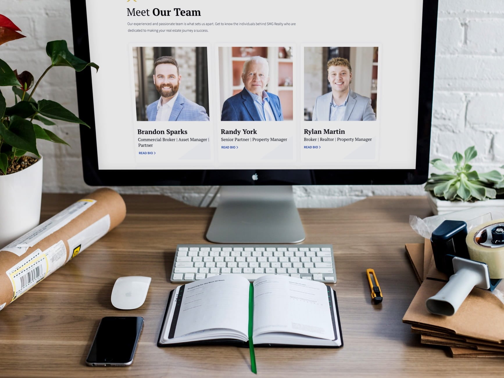 Desk with iMac showing SMG Realty team page, photos of Brandon Sparks, Randy York, Rylan Martin, notebook, supplies.