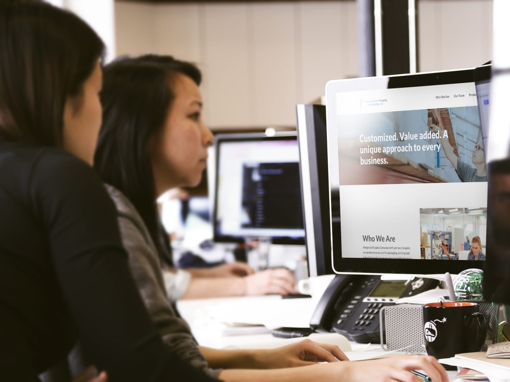 Two women at office desks looking at a monitor showing Integrated Supply Company website with 'Customized. Value added.' tagl