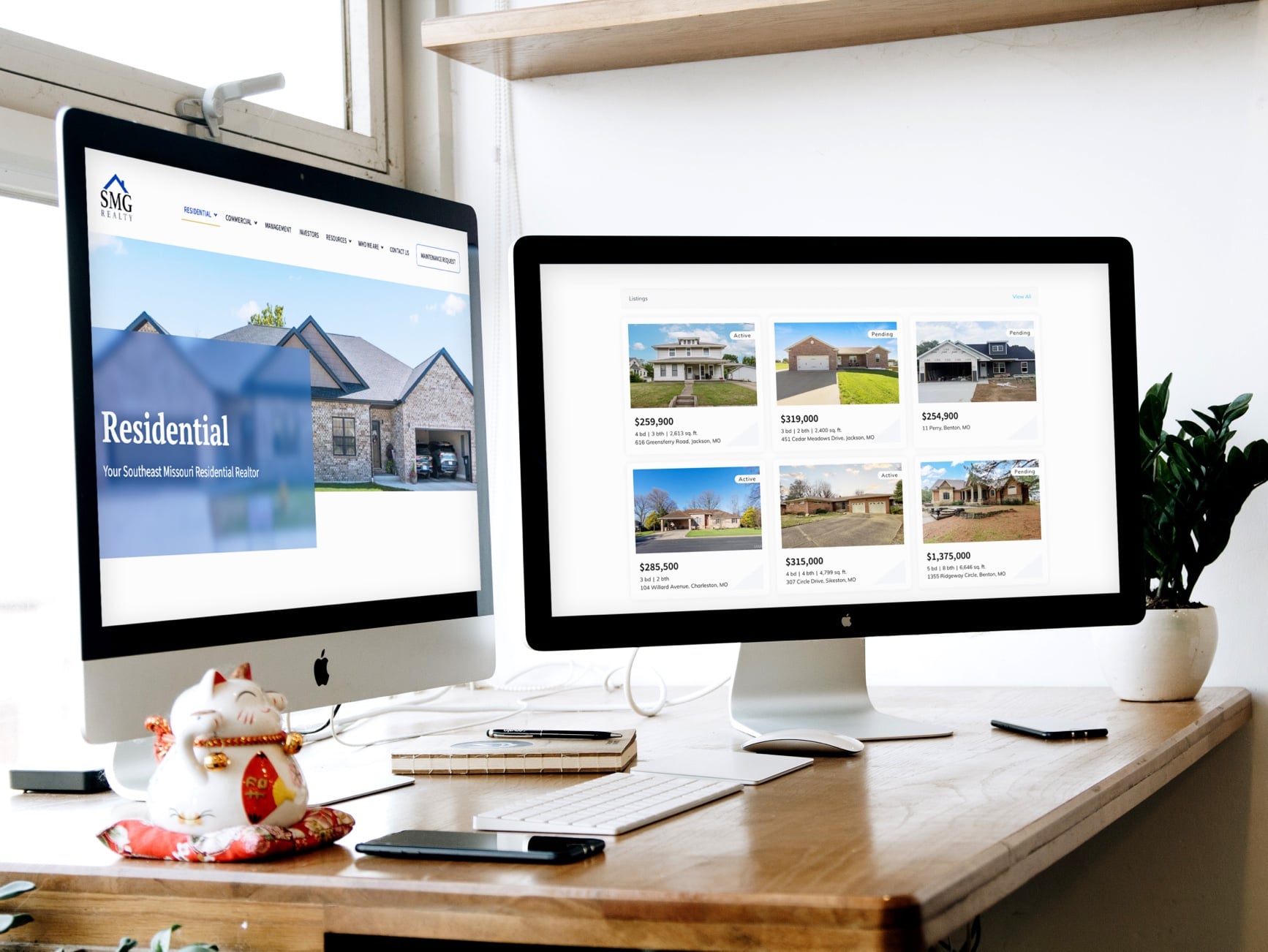 Two iMac monitors on a wooden desk displaying a residential real estate website with property listings and a home image.