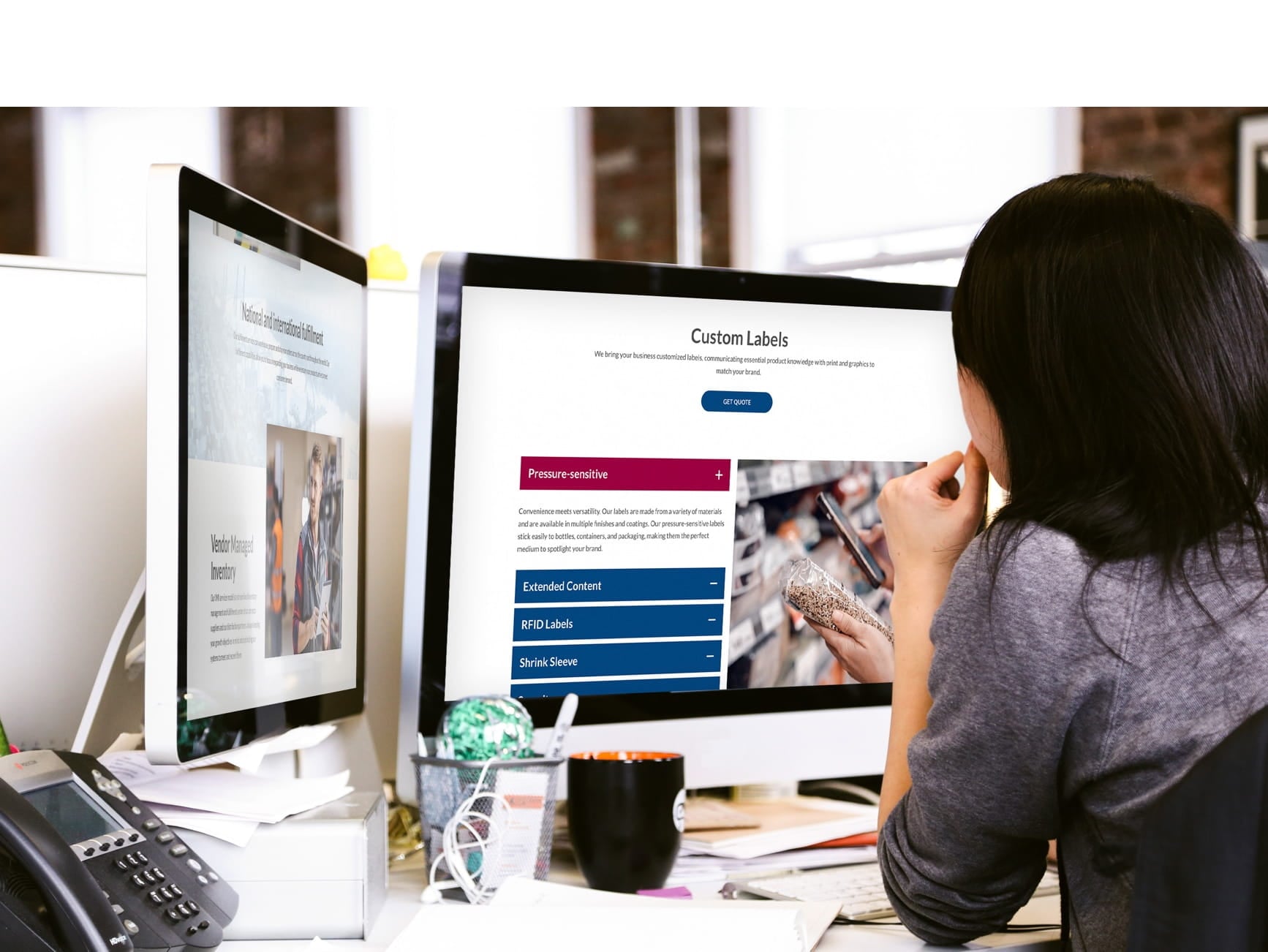 A woman sits at a desk, viewing a custom labels website on dual monitors in an office.