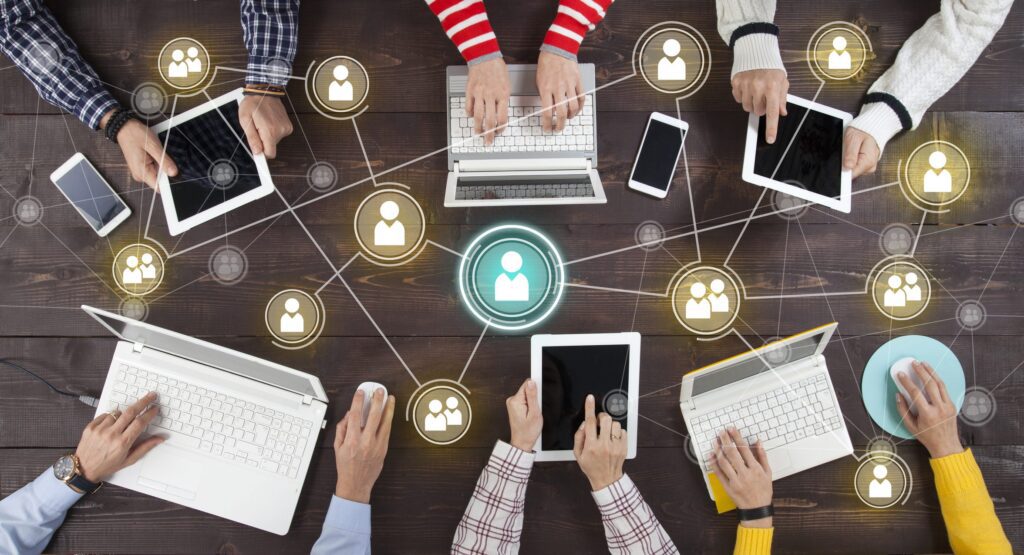 People at a dark table using laptops, tablets, phones, linked by glowing network icons for social media.