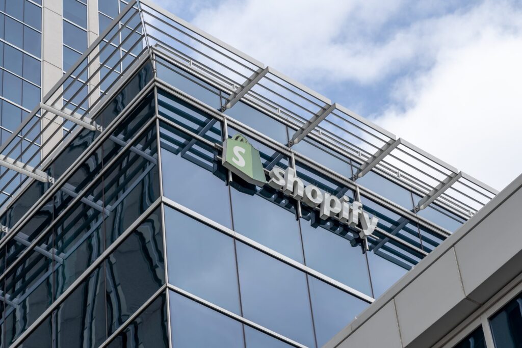 Shopify logo with green bag icon and white text on a glass building corner under blue sky