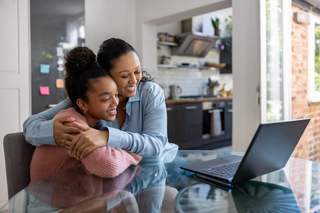 Mother and daughter hugging while smiling at a laptop on a glass table in a modern kitchen.