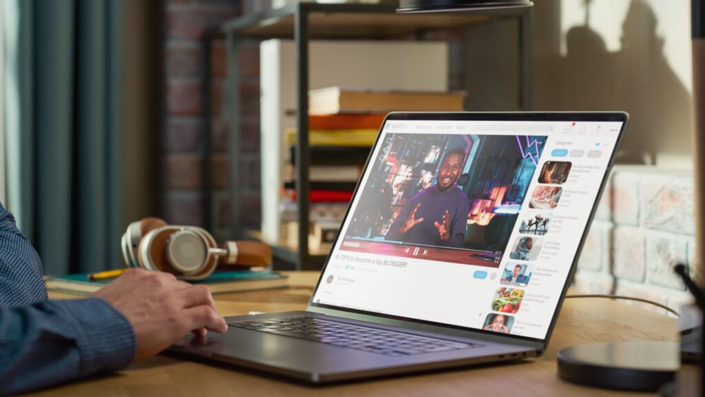 A person in a blue shirt uses a laptop to watch a video titled '10 Tips to become a top blogger' on a desk with headphones.