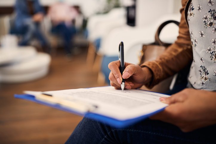 Person writing on a document with a pen while holding a blue clipboard in a waiting room.