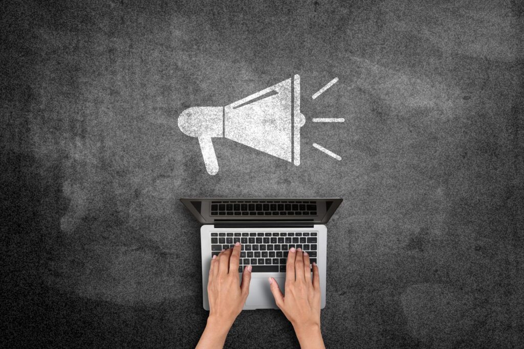Top-down view of hands typing on a silver laptop on a dark textured surface, with a white megaphone icon above.